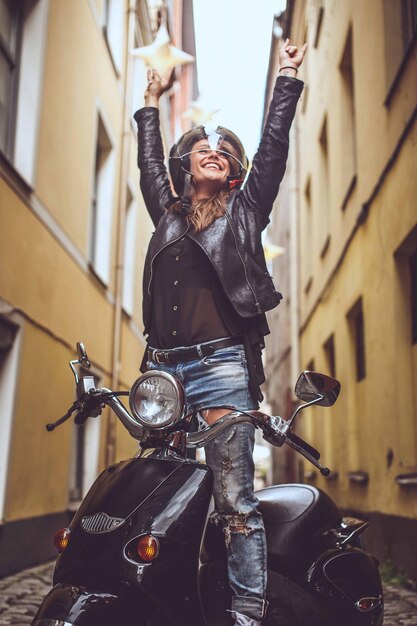 Young woman in blue jeans and black jacket standing on moto scooter with hands up.