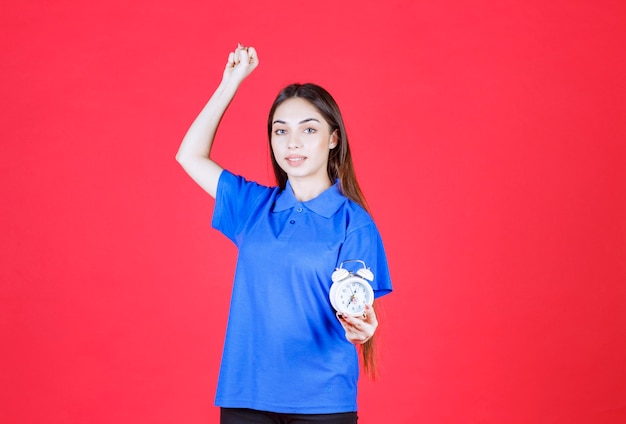 Young woman in blue shirt holding an alarm clock and showing positive hand sign