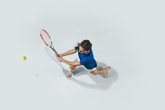 テニスをしている青いシャツを着た若い女性。白で隔離の屋内スタジオショット。上面図。