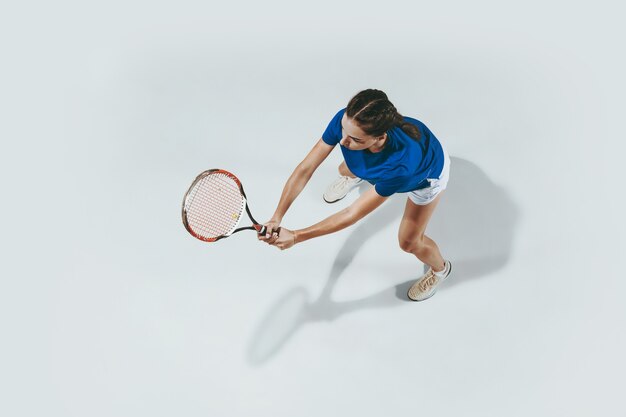 テニスをしている青いシャツの若い女性。彼女はラケットでボールを打つ。白で隔離された屋内ショット。若さ、柔軟性、パワー、そしてエネルギー。ネガティブスペース。上面図。
