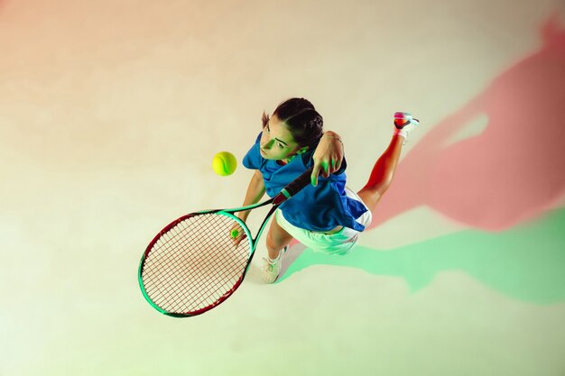 Young woman in blue shirt playing tennis. She hits the ball with a racket. Indoor  shot with mixed light. Youth, flexibility, power and energy. Top view.