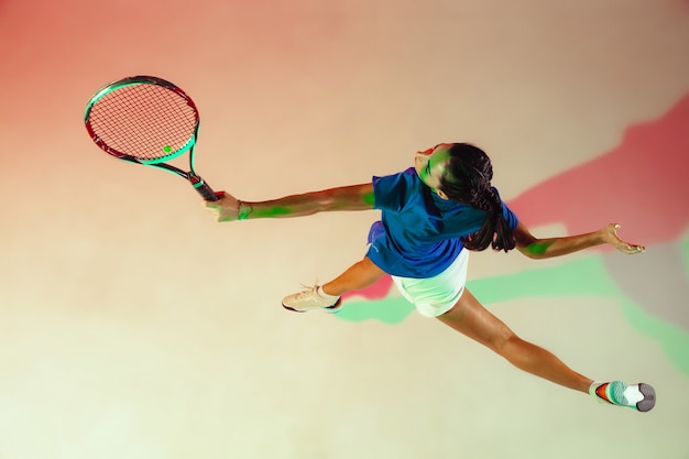 テニスをしている青いシャツの若い女性。彼女はラケットでボールを打つ。混合光による屋内撮影。若さ、柔軟性、パワー、そしてエネルギー。上面図。
