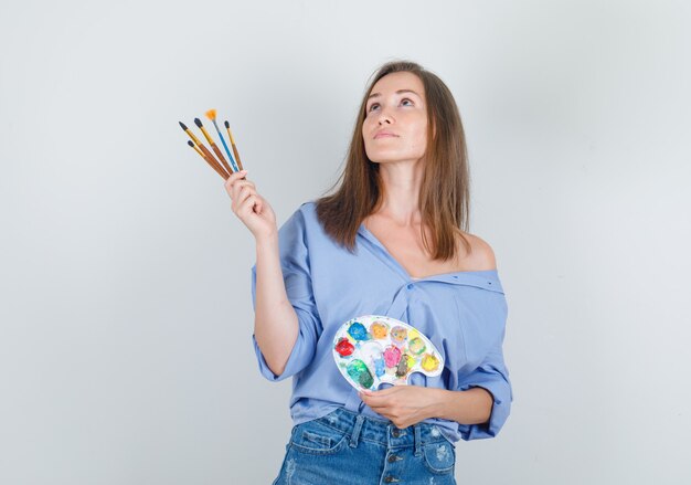 Young woman in blue shirt, shorts holding painting tools and looking hopeful