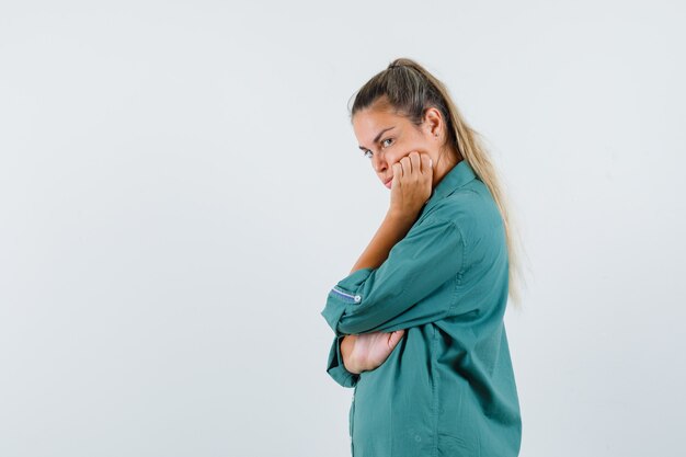 Young woman in blue shirt standing with hand on jaw and looking dissatisfied .