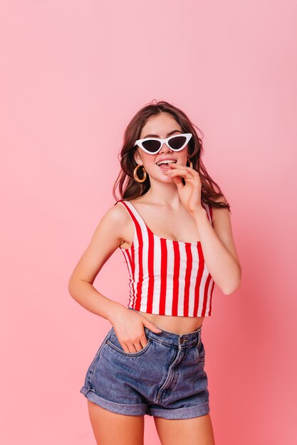 Young woman in blue shorts and red-white top coquettishly posing on pink wall