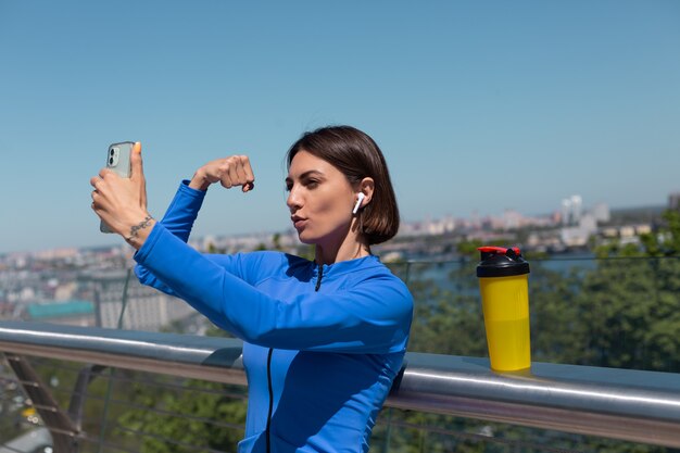 Молодая женщина в синей спортивной одежде на мосту жарким солнечным утром с беспроводными наушниками и мобильным телефоном, делает селфи, фото-видео для социальных сетей, показывает свои мышцы бицепса