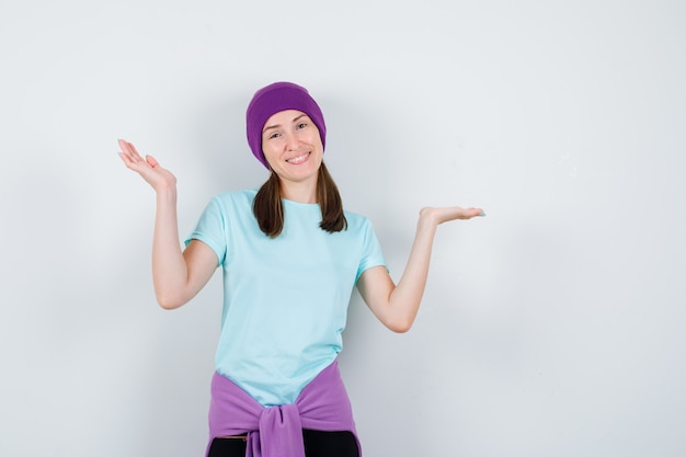 Foto gratuita giovane donna in maglietta blu, berretto viola che allarga le mani con gioia e sembra allegra, vista frontale.