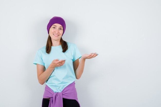 Young woman in blue t-shirt, purple beanie stretching hands as displaying something and looking cheery , front view.