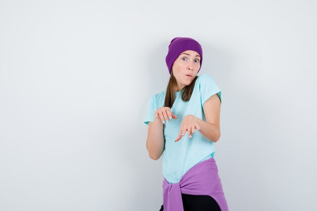 Young woman in blue t-shirt, purple beanie stretching hands toward camera and looking surprised , front view.