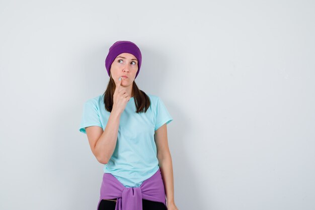Молодая женщина в синей футболке, фиолетовой шапочке с указательным пальцем на подбородке, думающая о чем-то и задумчивая, вид спереди.