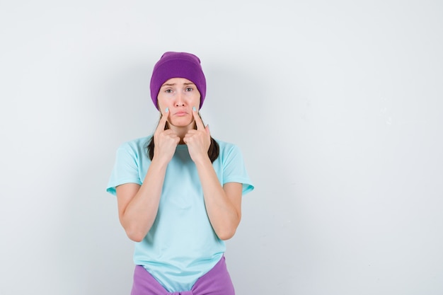Young woman in blue t-shirt, purple beanie withindex fingers near mouth, curving lips and looking dismal , front view.