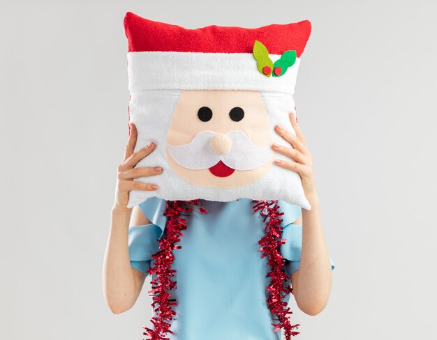 Young woman in blue top holding christmas pillow in front of her face hiding her face 