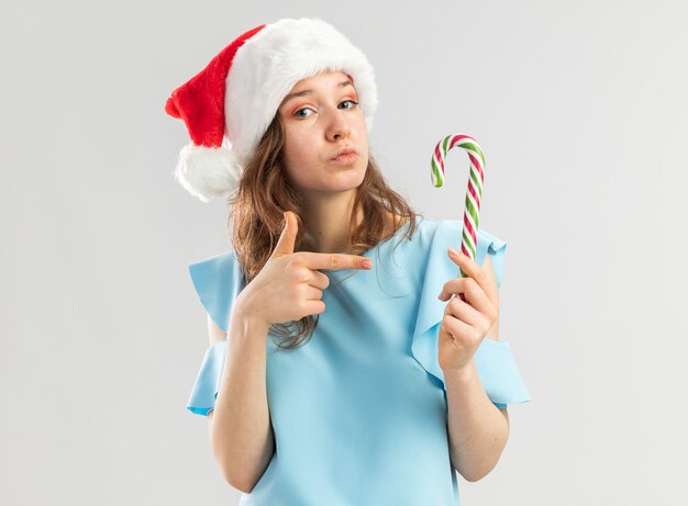 Young woman in blue top and santa hat holding candy cane pointng with index finger at it looking confident 