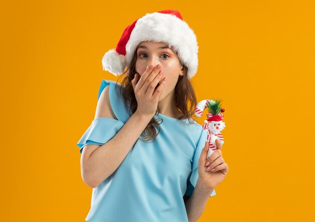 Молодая женщина в синем топе и шляпе санта-клауса, держащая рождественскую конфету, выглядит шокированной, прикрывая рот рукой