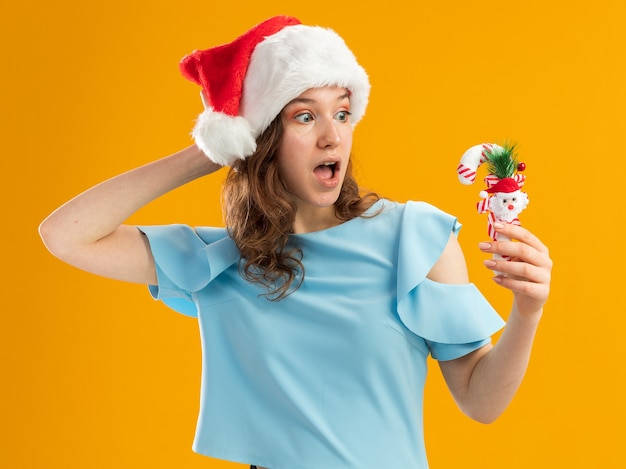 Молодая женщина в синем топе и шляпе санта-клауса держит рождественскую конфету, глядя на нее, изумленно положив руку на голову