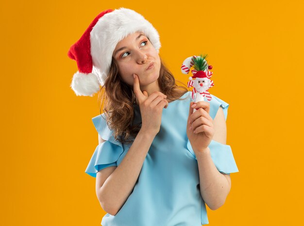 Young woman in blue top and santa hat holding christmas candy cane looking up puzzled 