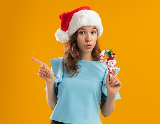 Young woman in blue top and santa hat holding christmas candy cane looking worried pointing with index finger to the side