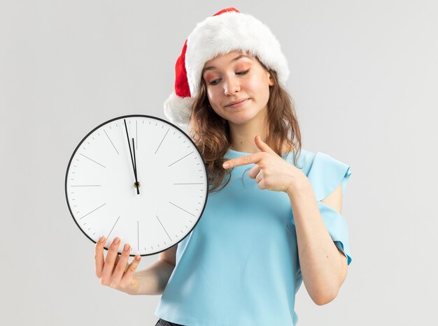 Young woman in blue top and santa hat holding wall clock pointing with index finger at it smiling confident 