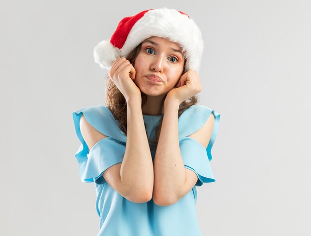 Young woman in blue top and santa hat looking confused making wry mouth with disappointed expression 