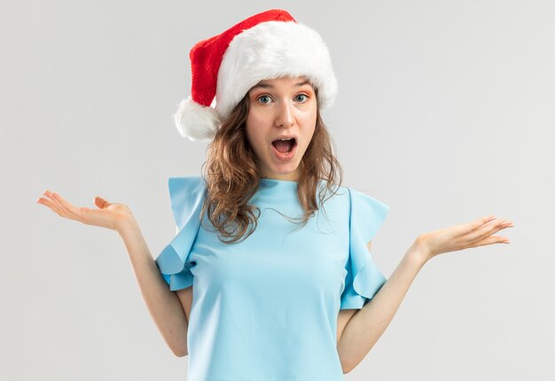 Молодая женщина в синем топе и новогодней шапке выглядит смущенной и удивленной с поднятыми руками