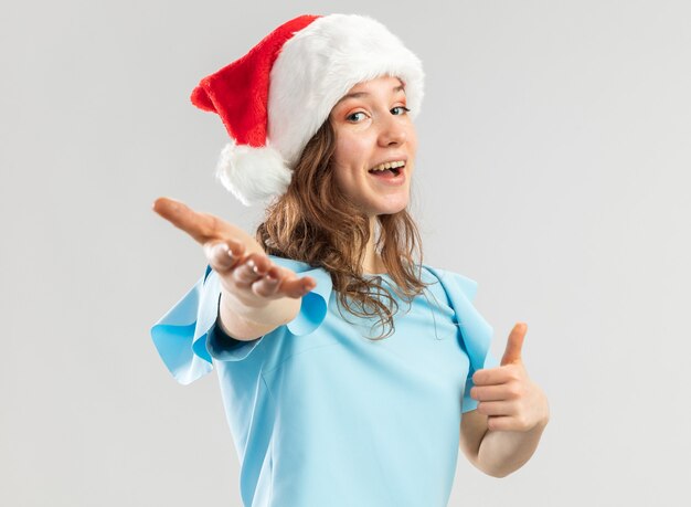 Young woman in blue top and santa hat looking happy and positive making come here gesture with hand 