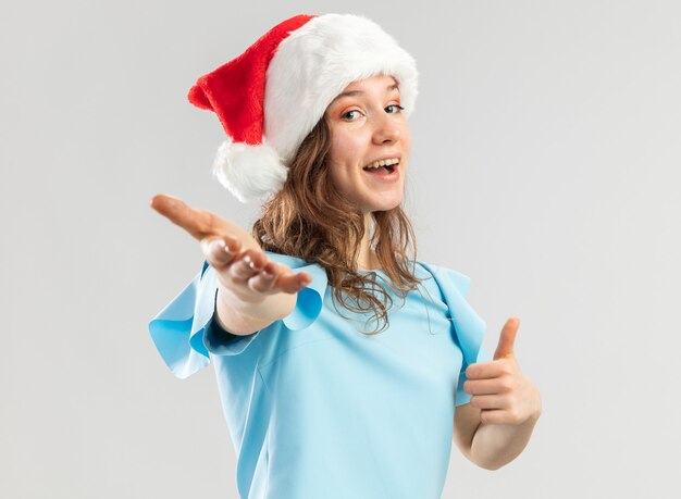 파란색 상단과 산타 모자에 젊은 여자가 행복하고 긍정적 인 만들기를 찾고 여기 손으로 제스처