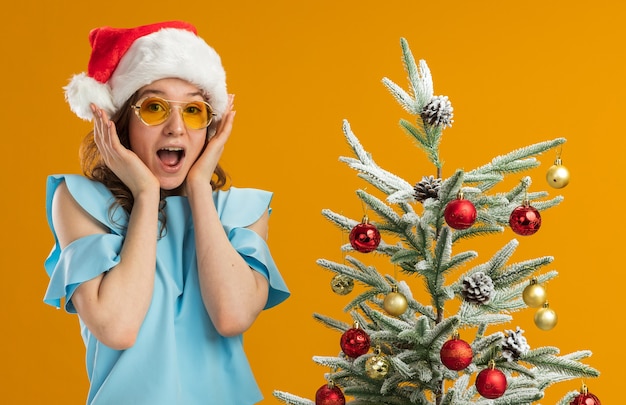 Young woman in blue top and santa hat wearing yellow glasses   amazed and surprsied standing next to a christmas tree over orange wall