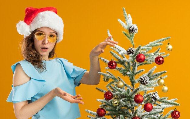 오렌지 배경 위에 서 혼란 스 러 워 찾고 크리스마스 트리를 장식하는 노란색 안경을 쓰고 파란색 위쪽 및 산타 모자에 젊은 여자