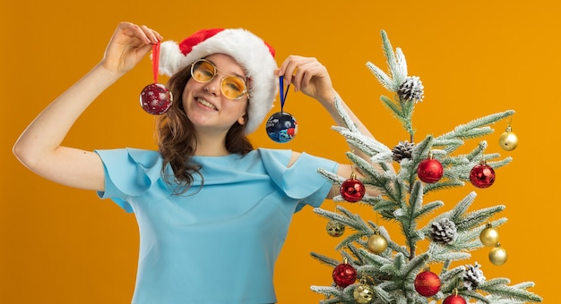 Young woman in blue top and santa hat wearing yellow glasses holding christmas balls looking at camera happy and cheerful standing next to a christmas tree over orange background