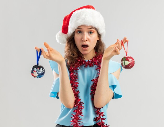 Young woman in blue top and santa hat with tinsel around her neck holding christmas balls looking confused and surprised 