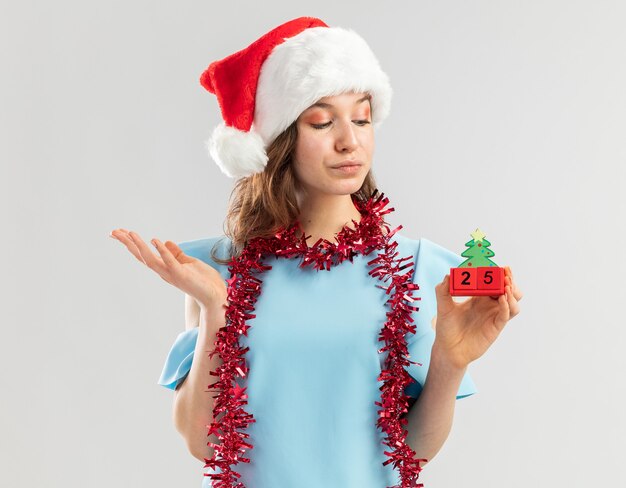 Молодая женщина в синем топе и шляпе санта-клауса с мишурой на шее держит игрушечные кубики с рождественским свиданием, уверенно выглядит с поднятой рукой