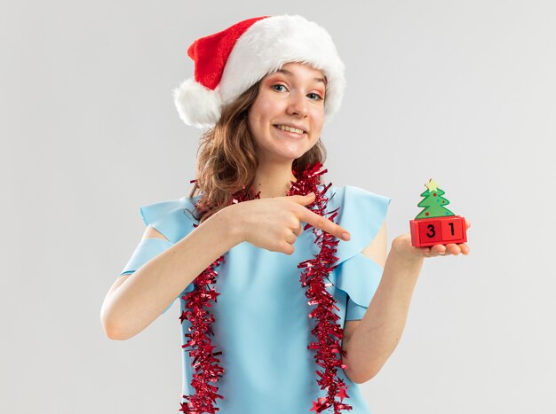 Молодая женщина в синем топе и новогодней шапке с мишурой на шее держит игрушечные кубики с новогодней датой, указывая указательным пальцем на кубики, счастливая и веселая