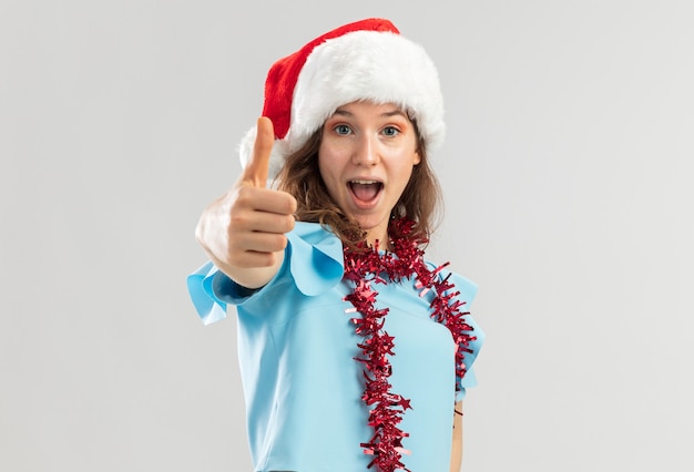 Young woman in blue top and santa hat with tinsel around her neck looking happy and excited showing thumbs up 