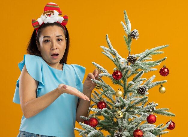 面白いクリスマスの縁を身に着けている青いトップの若い女性は、オレンジ色の背景の上に立って不満で腕を上げて混乱しているように見えるクリスマスツリーを飾る