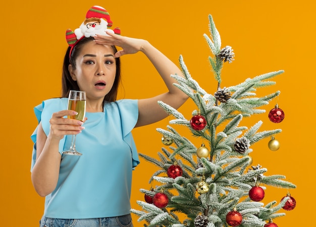 Young woman in  blue top wearing funny christmas  rim on head holding glass of champagne looking far away being surprised standing next to a  christmas tree  over orange background