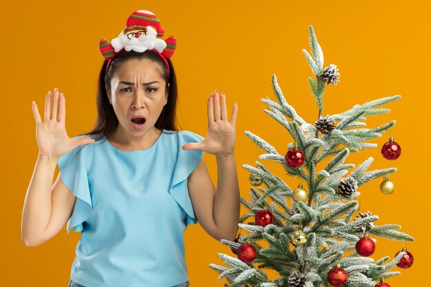 オレンジ色の背景の上のクリスマスツリーの横に立っている腕を上げて叫んで怒っている顔でカメラを見て頭に面白いクリスマスの縁を身に着けている青いトップの若い女性