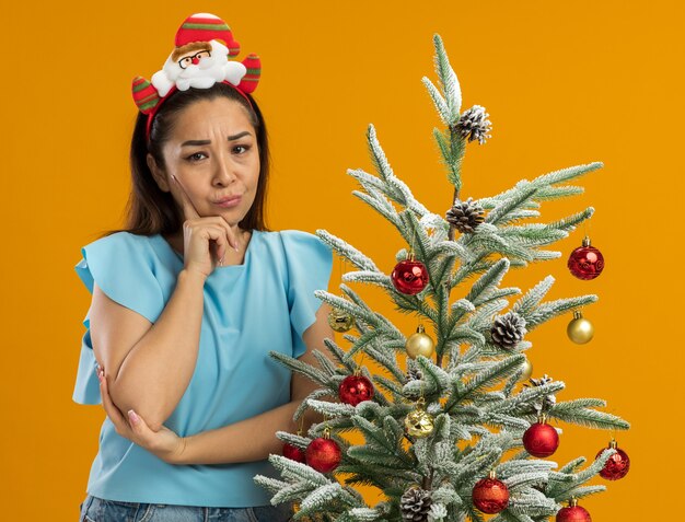 Young woman in  blue top wearing funny christmas  rim on head looking at camera with skeptic expression standing next to a  christmas tree over orange background