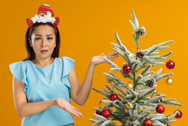 頭に面白いクリスマスの縁を身に着けている青いトップの若い女性は、オレンジ色の背景に不快感と憤慨の手を上げて混乱しているように見えるクリスマスツリーの横に立っています