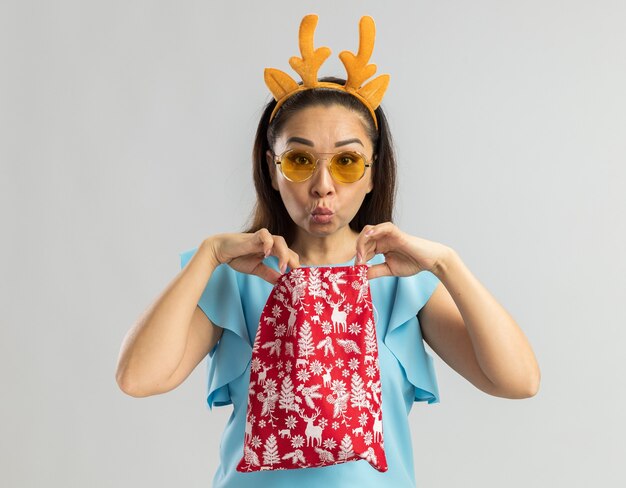 Young woman in blue top wearing funny rim with deer horns and yellow glasses holding christmas red bag opening it looking intrigued 