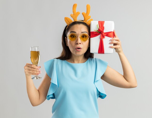 Young woman in blue top wearing funny rim with deer horns and yellow glasses holding glass of champagne and christmas present looking surprised and worried 