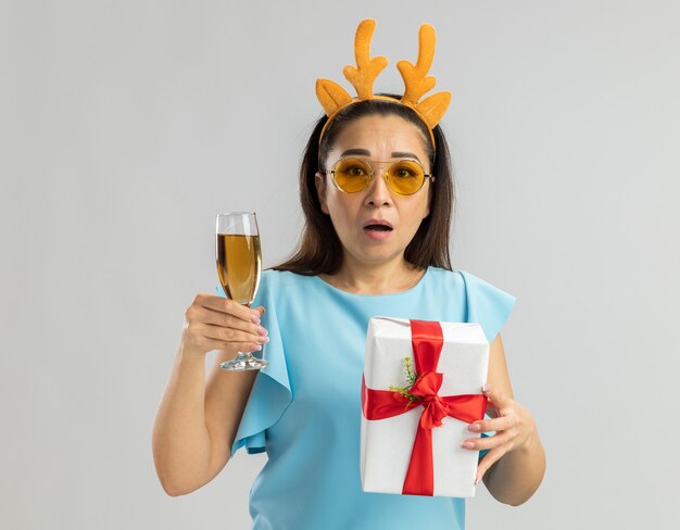 Young woman in blue top wearing funny rim with deer horns and yellow glasses holding glass of champagne and christmas present looking worried and confused 