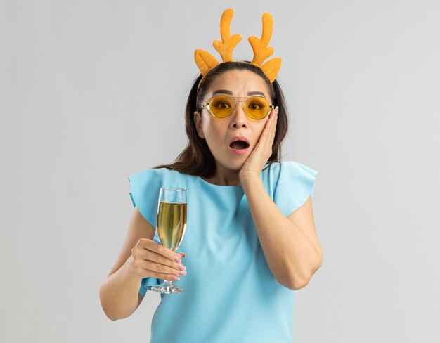 Young woman in blue top wearing funny rim with deer horns and yellow glasses holding glass of champagne looking amazed and surprised 