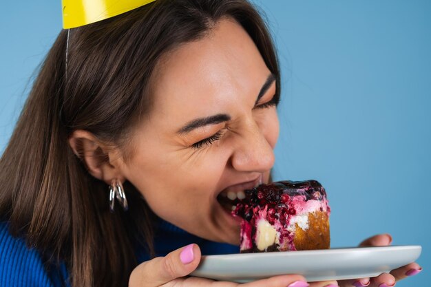 Young woman on a blue wall celebrates a birthday, holds a piece of cake, happy, bites the cake