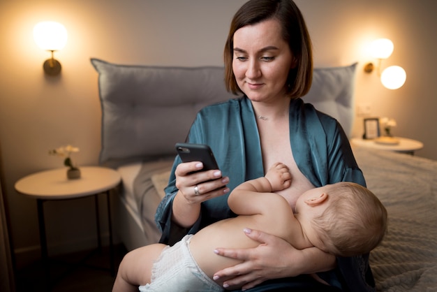 Giovane donna che allatta al seno il suo bambino carino mentre controlla il suo telefono