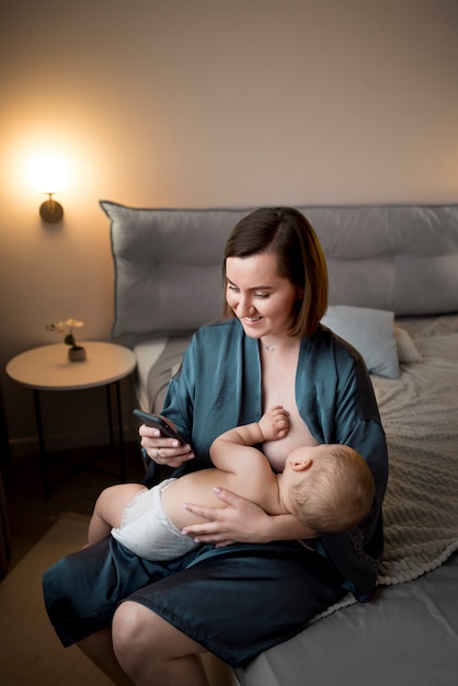 Giovane donna che allatta al seno il suo bambino carino mentre controlla il suo telefono