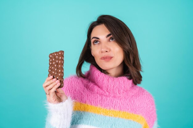 A young woman in a bright multi-colored sweater on blue enjoys every piece of a delicious bar of airy milk chocolate