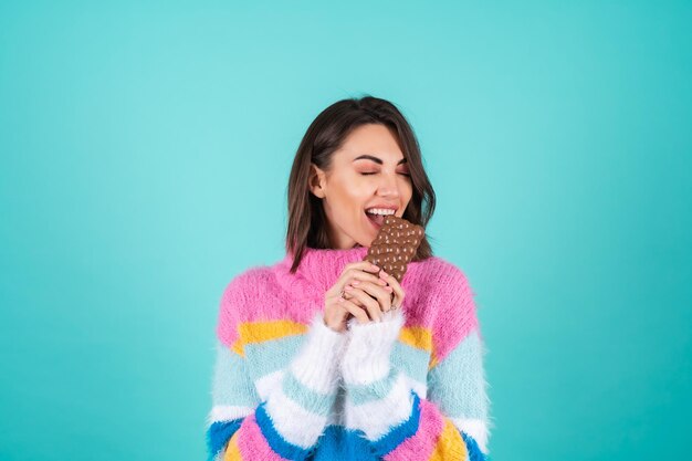 A young woman in a bright multi-colored sweater on blue enjoys every piece of a delicious bar of airy milk chocolate