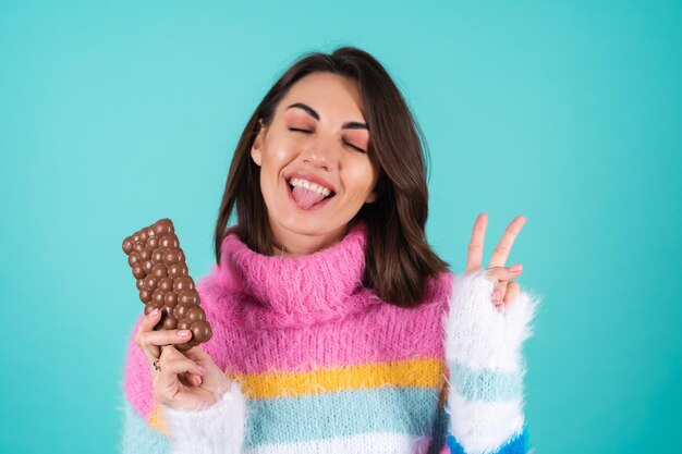 A young woman in a bright multi-colored sweater on blue enjoys every piece of a delicious bar of airy milk chocolate