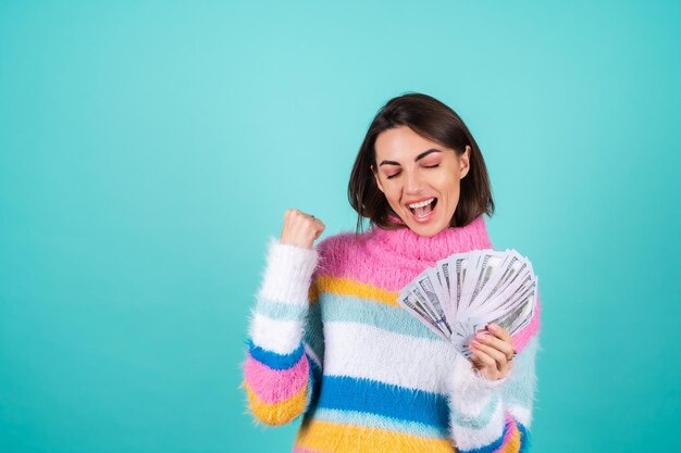 A young woman in a bright multicolored sweater on blue holds a bundle of dollars