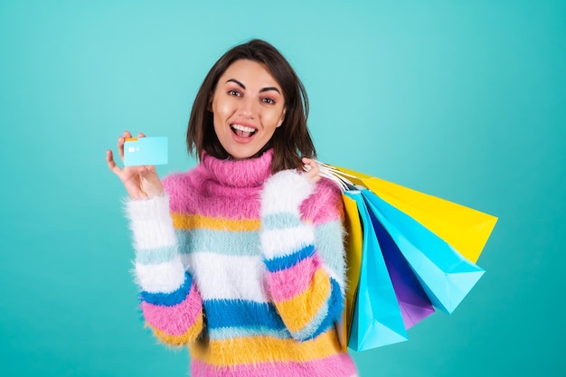 Young woman in a bright multicolored sweater on blue holds a credit card and shopping bags